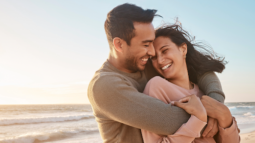 Man and woman laughing and hugging while enjoying a seaside sunset.